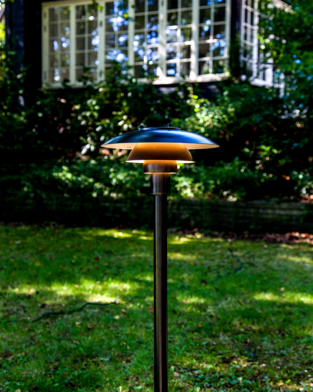 A dark-finished PH 3-2½ Bollard stands on a lawn in front of a house, lighting the garden. © 2026 Louis Poulsen.