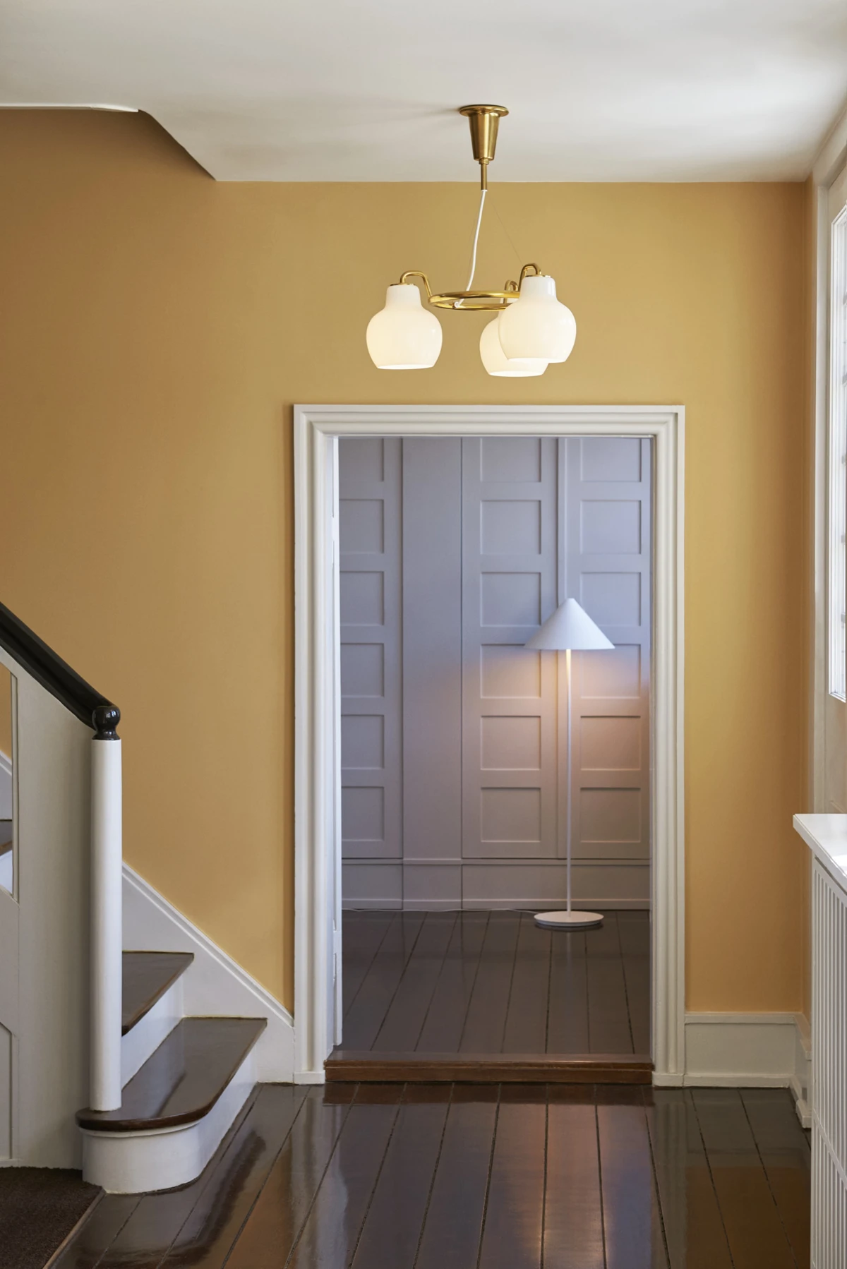 Een VL Ring Crown 3-5-7 hanglamp met opaalglazen kappen en een witte Keglen vloerlamp in een okergele hal. © 2026 Louis Poulsen.