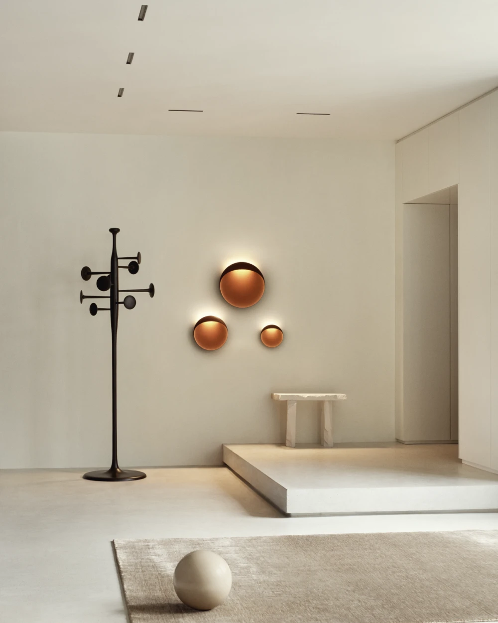 Three Flindt Wall Lamps cast a warm glow on the wall of a minimalist hallway with pale stone surfaces. © 2026 Louis Poulsen.