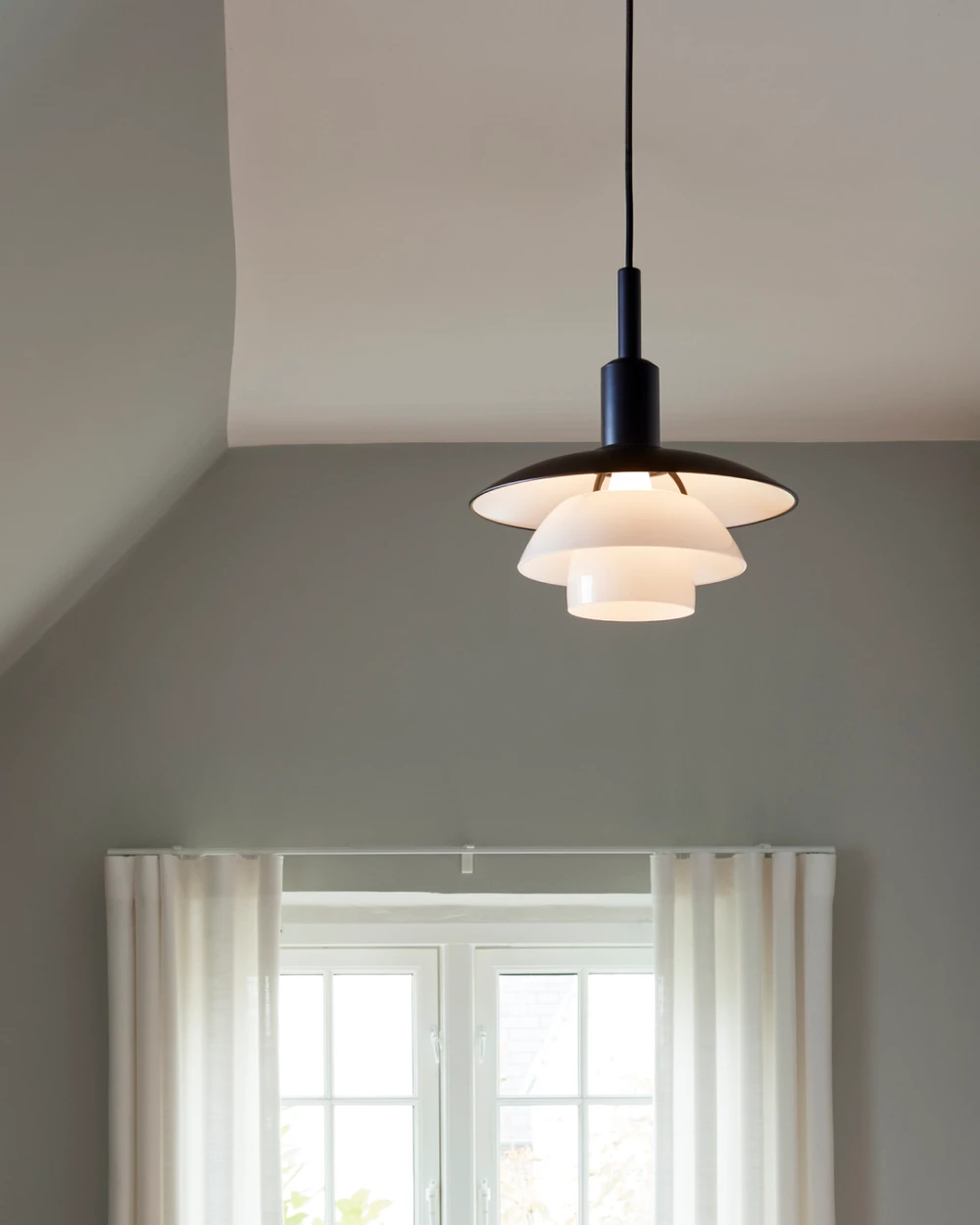 A black PH 3/3 Pendant with opal glass shades lights the area above a window in a quiet hallway. © 2026 Louis Poulsen.