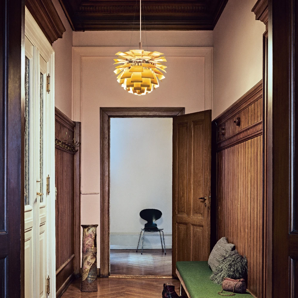 A golden PH Artichoke pendant lights a hallway with dark wood panelling and a green bench. © 2026 Louis Poulsen.