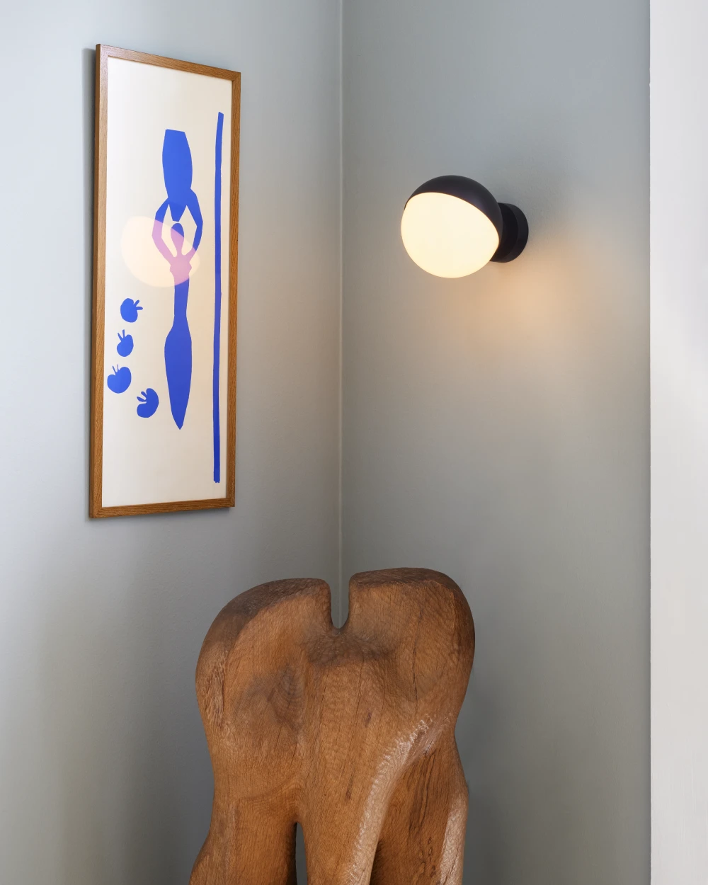 A black VL Studio Wall Lamp with an opal-white shade glows in a hallway beside framed art and a wooden sculpture. © 2026 Louis Poulsen.