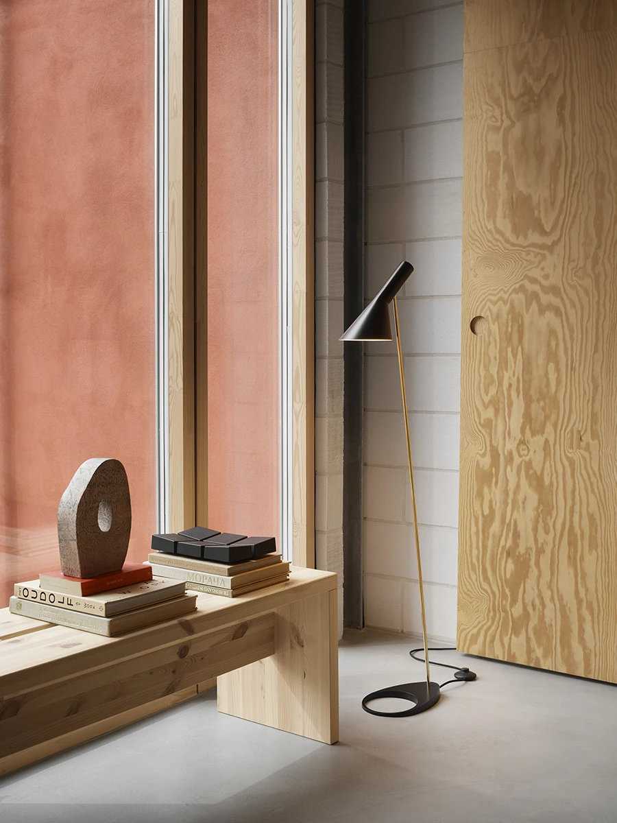 A black AJ Floor Lamp with a brass-coloured stem stands beside a light wood bench in a hallway. © 2026 Louis Poulsen.