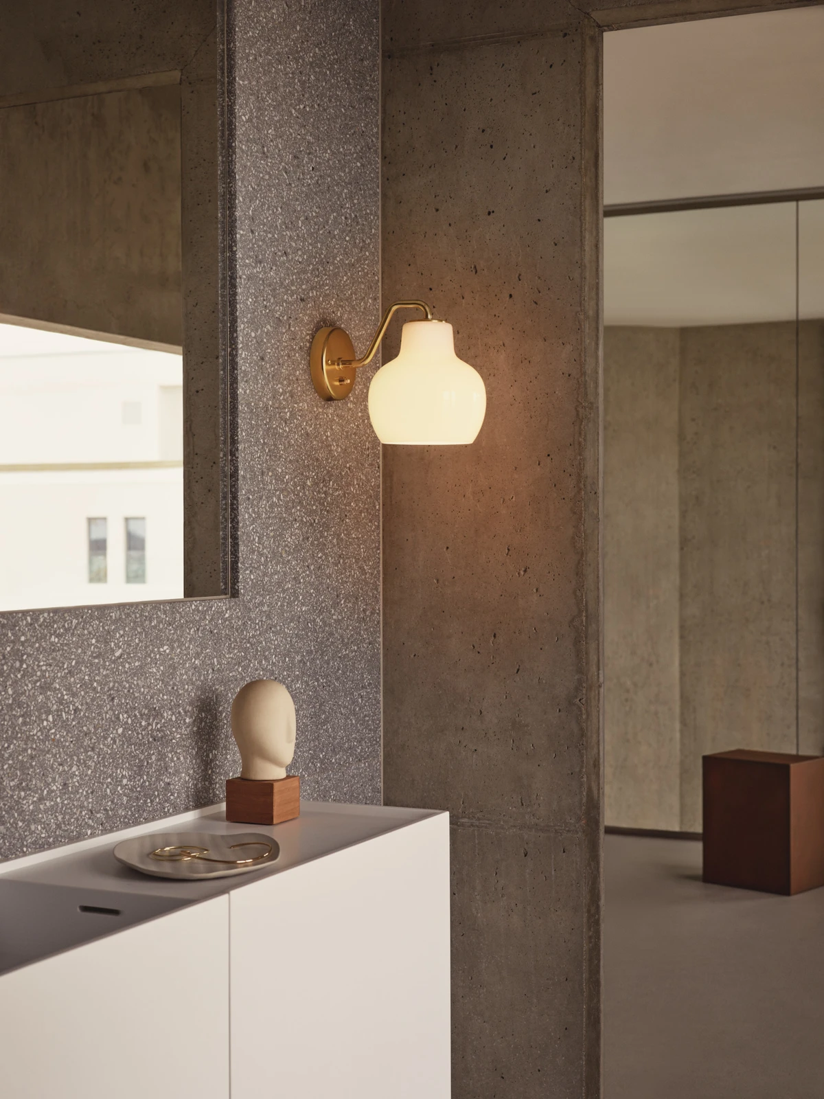 A brass-finish VL Ring Crown 1 Wall Lamp with opal glass casts a warm glow in a hallway with concrete walls. © 2026 Louis Poulsen.