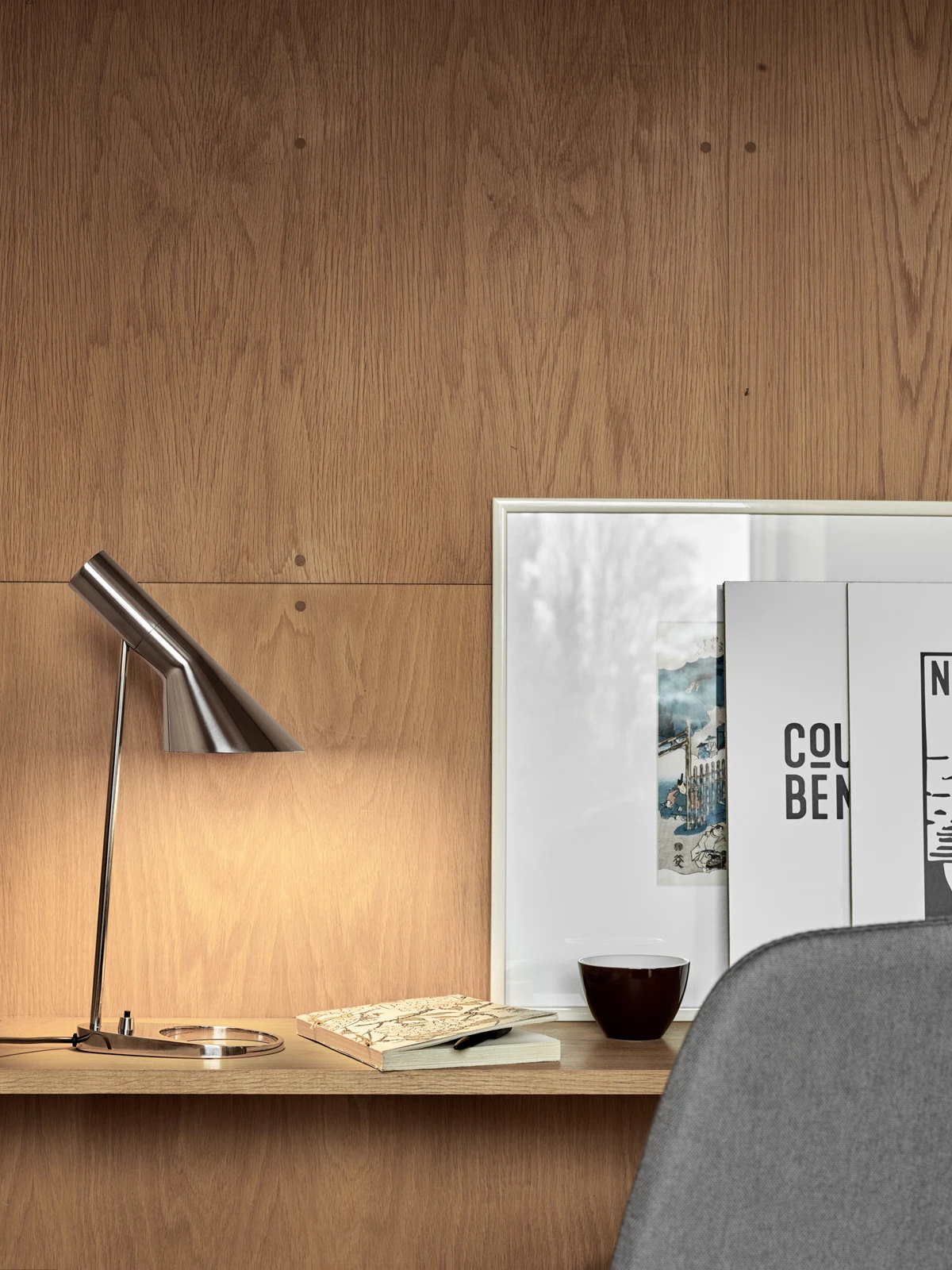 De AJ tafellamp van roestvrij staal verlicht een bureau tegen een houten wand. © 2026 Louis Poulsen.