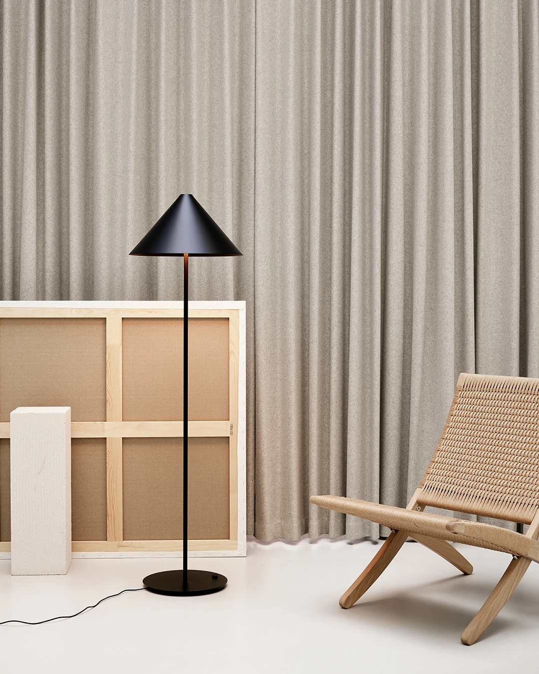 A black Keglen Floor Lamp stands before grey curtains beside a canvas and a woven light-wood lounge chair. © 2026 Louis Poulsen.
