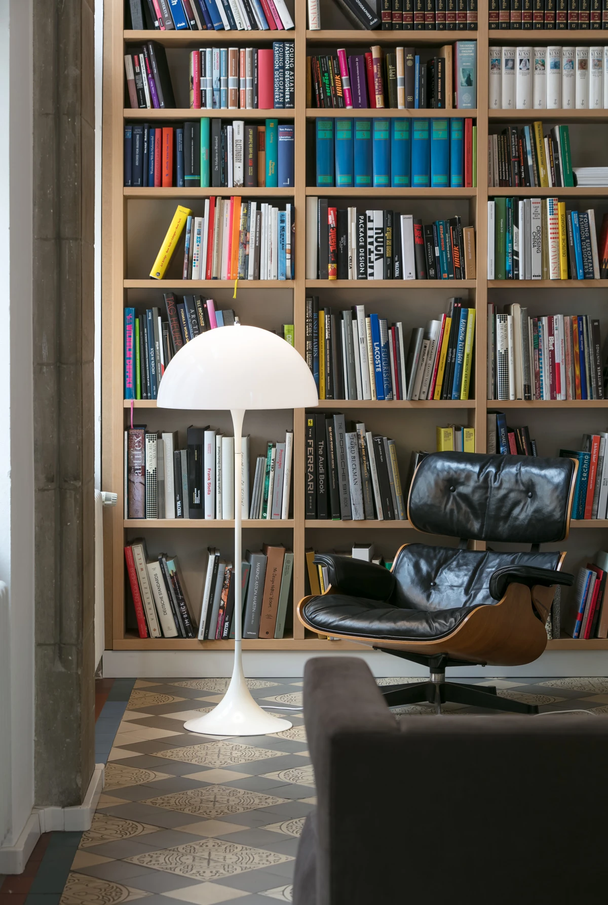 A white Panthella Floor Lamp stands before a bookcase beside a black leather chair on patterned tile flooring. © 2026 Louis Poulsen.