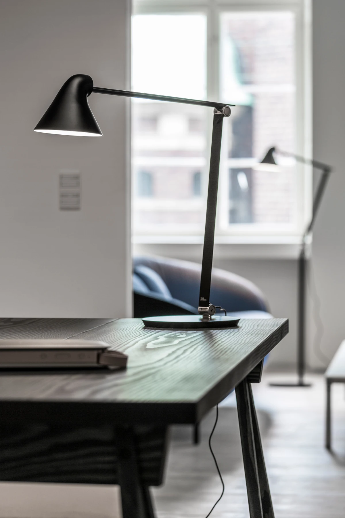 A black AJ Table Lamp sits on a dark wooden desk in a bright home office. © 2026 Louis Poulsen.