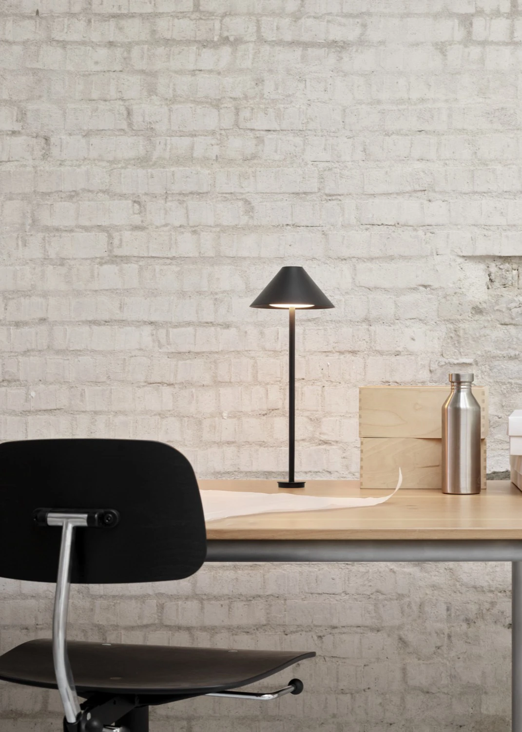 A black Keglen Table Lamp illuminates a desk against a white brick wall. © 2026 Louis Poulsen.