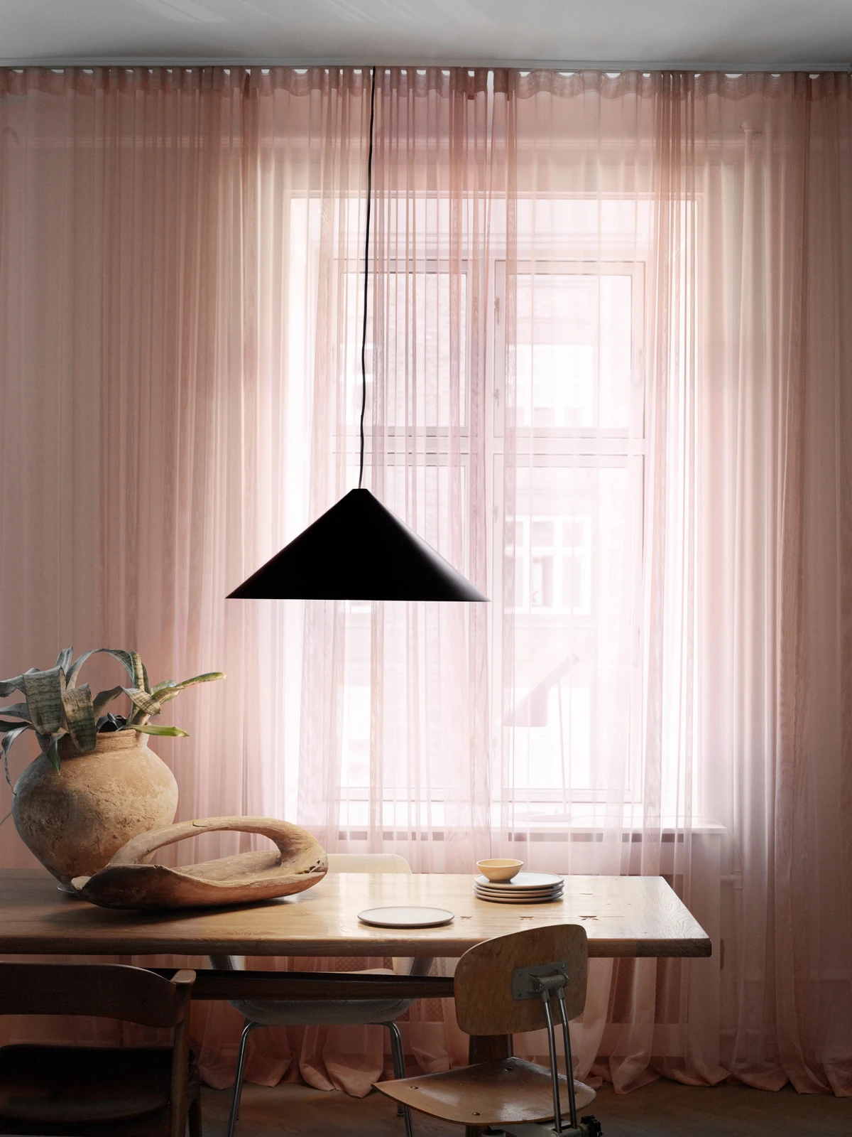 Een zwarte Keglen-hanglamp hangt boven een houten tafel voor doorschijnende roze gordijnen. © 2026 Louis Poulsen.