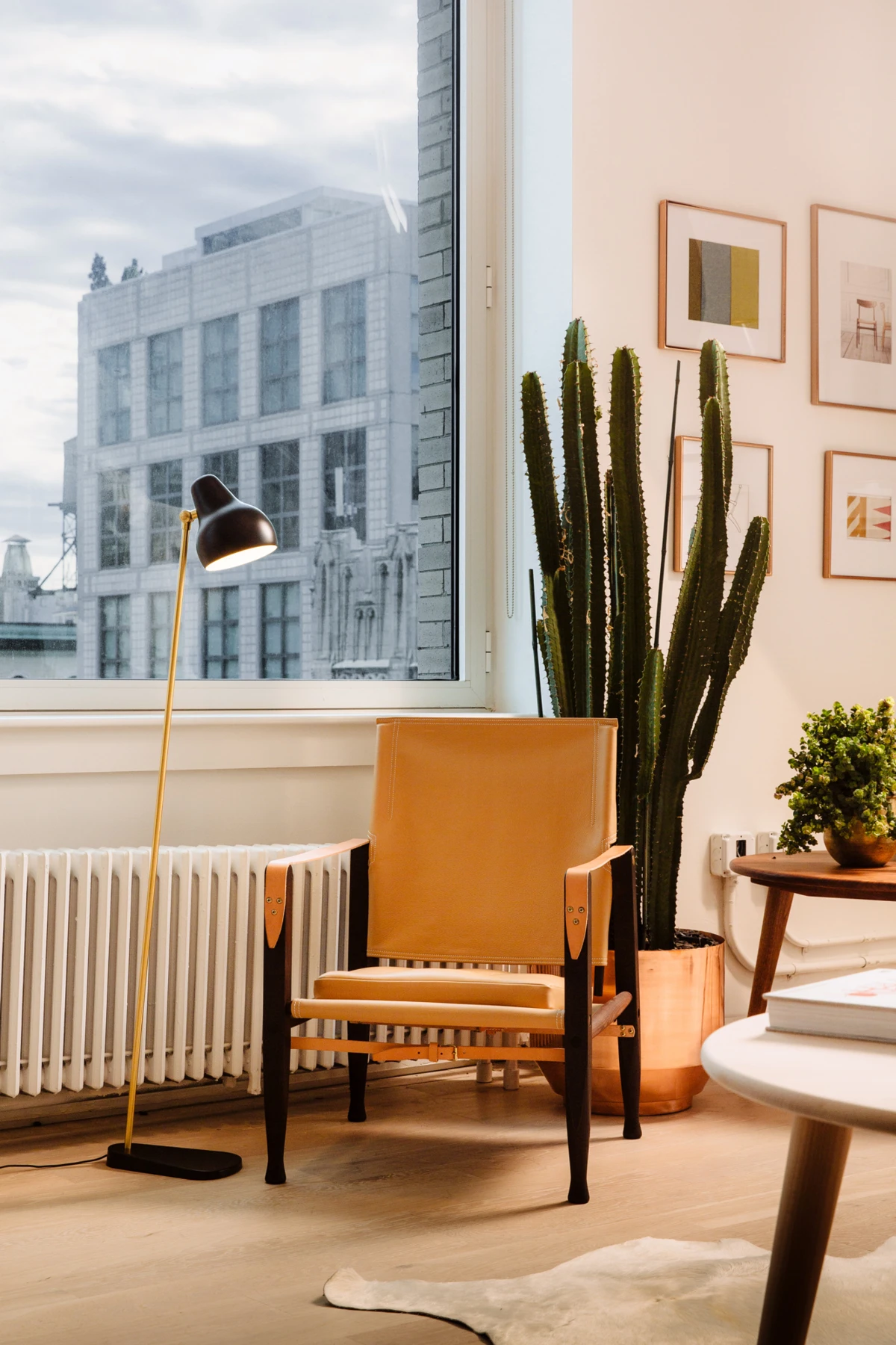 Een AJ Floor Lamp met zwarte kap en messingkleurige staander staat naast een leren fauteuil in een lichte ruimte met grote ramen. © 2026 Louis Poulsen.