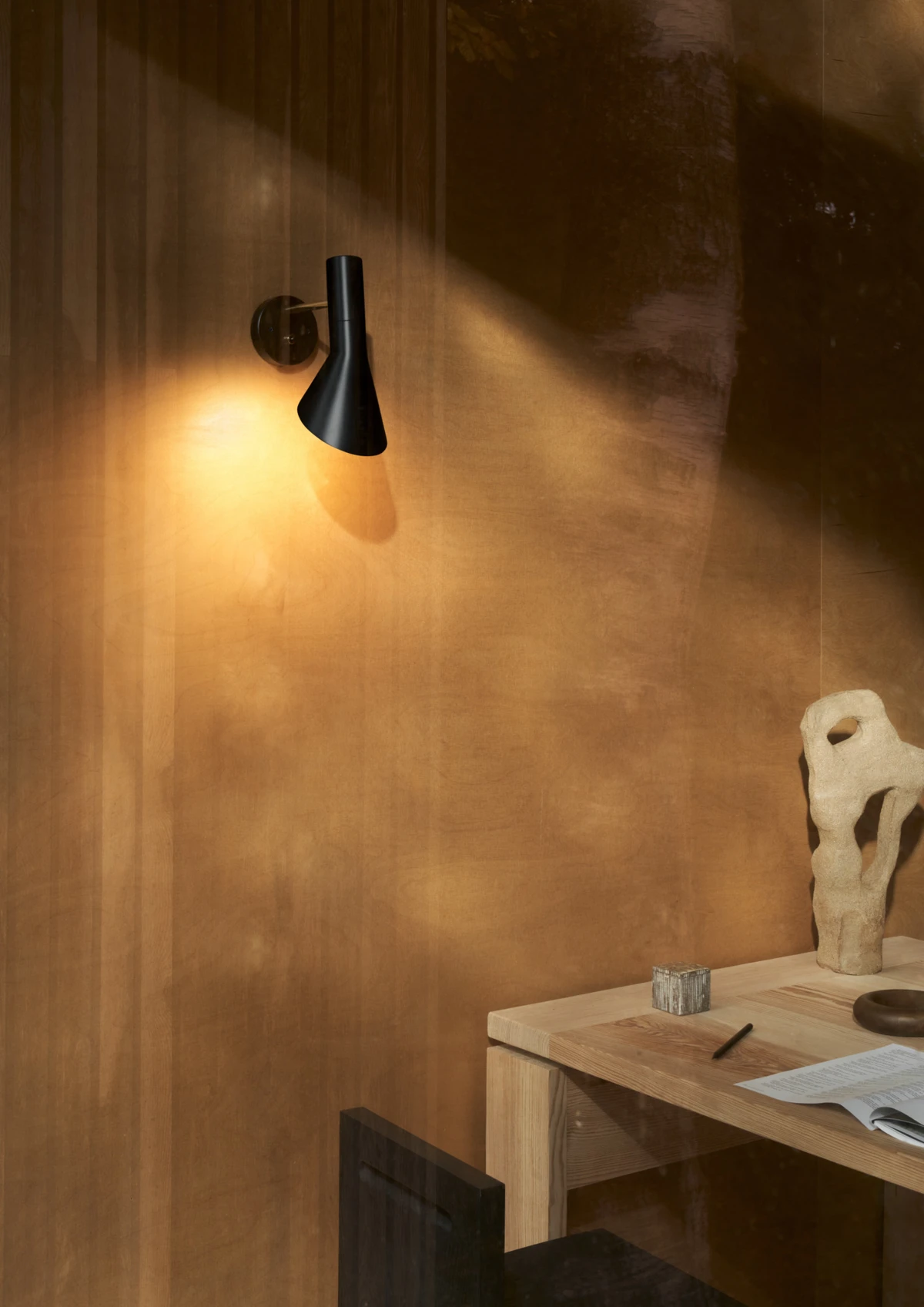 A black AJ Wall Lamp with a brass-finish mount casts warm light over a pale wooden desk. © 2026 Louis Poulsen.
