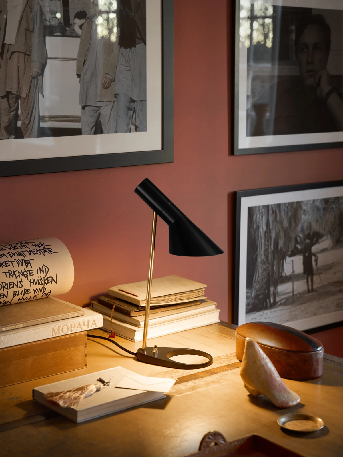 A black AJ Table Lamp with a brass stem illuminates a wooden desk surrounded by books and framed photographs. © 2026 Louis Poulsen.