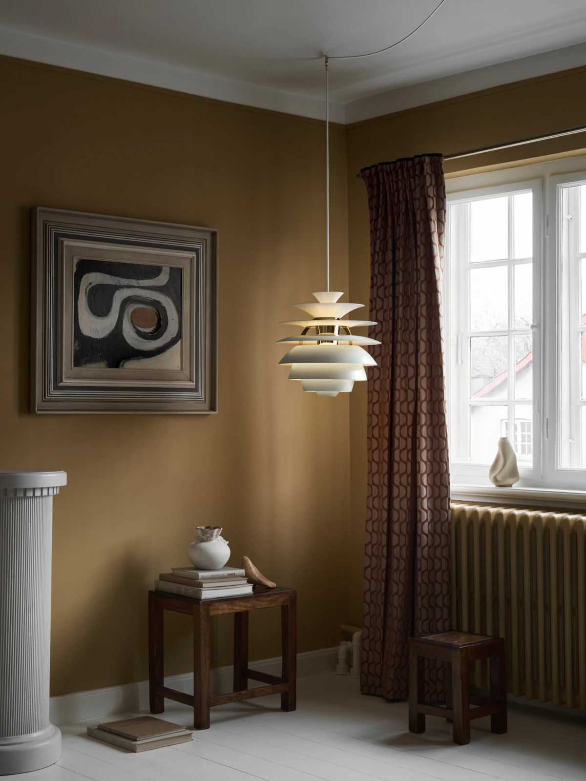 Een witte PH Snowball hanglamp hangt in een warm, aards getinte kamer naast een groot raam. © 2026 Louis Poulsen.