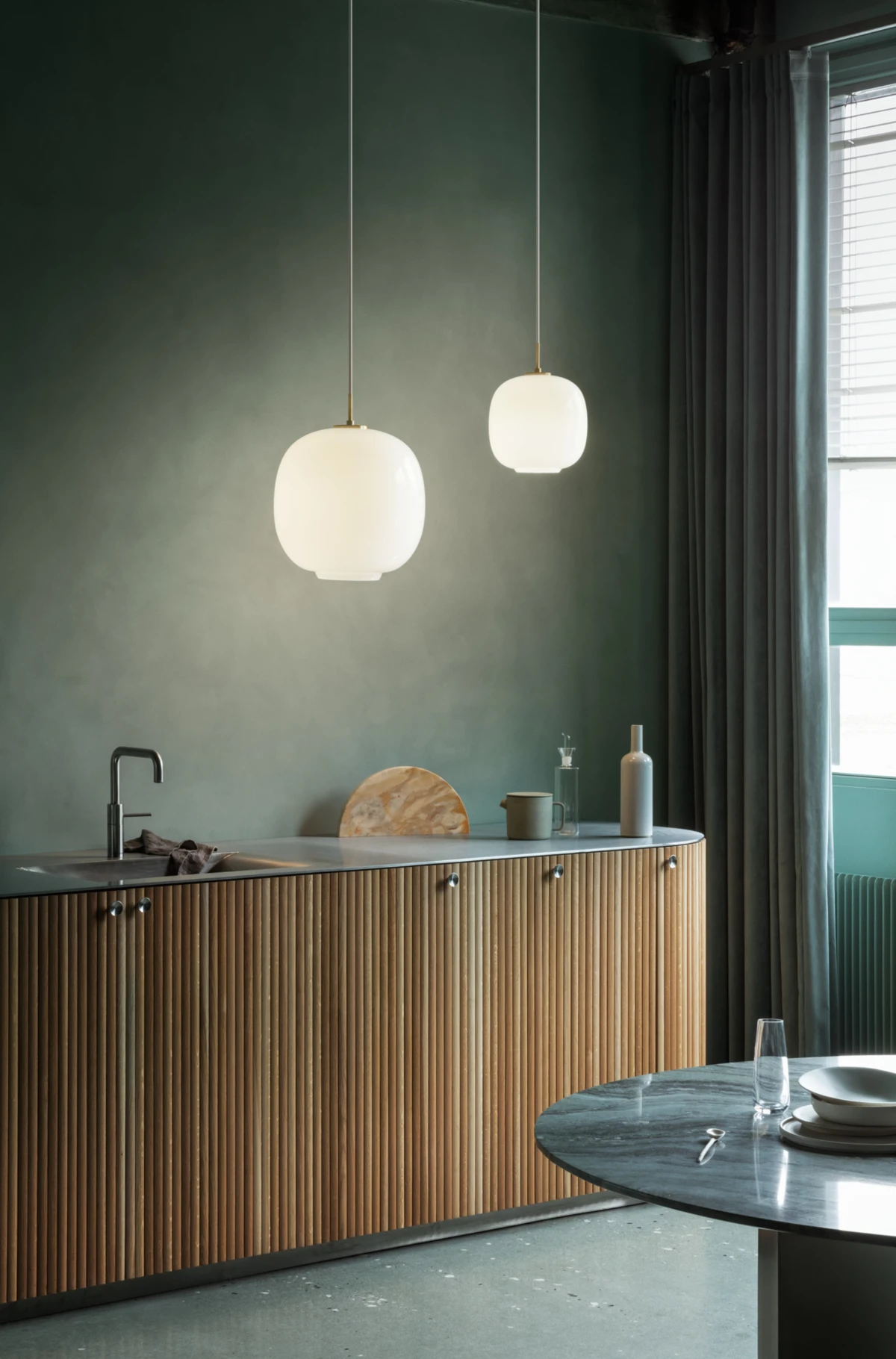 Two opal white Wohlert Pendants hang above a wood-ribbed kitchen island against a dark green wall. © 2026 Louis Poulsen.