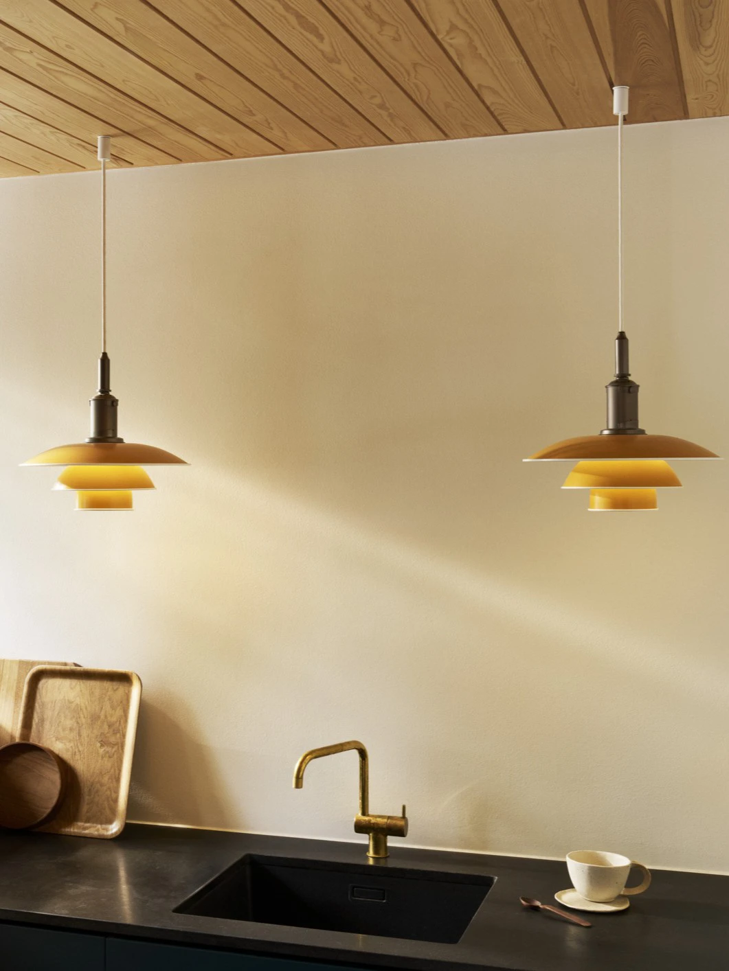Two yellow PH 3½-3 pendants hang above a dark stone kitchen counter with a brass tap. © 2026 Louis Poulsen.