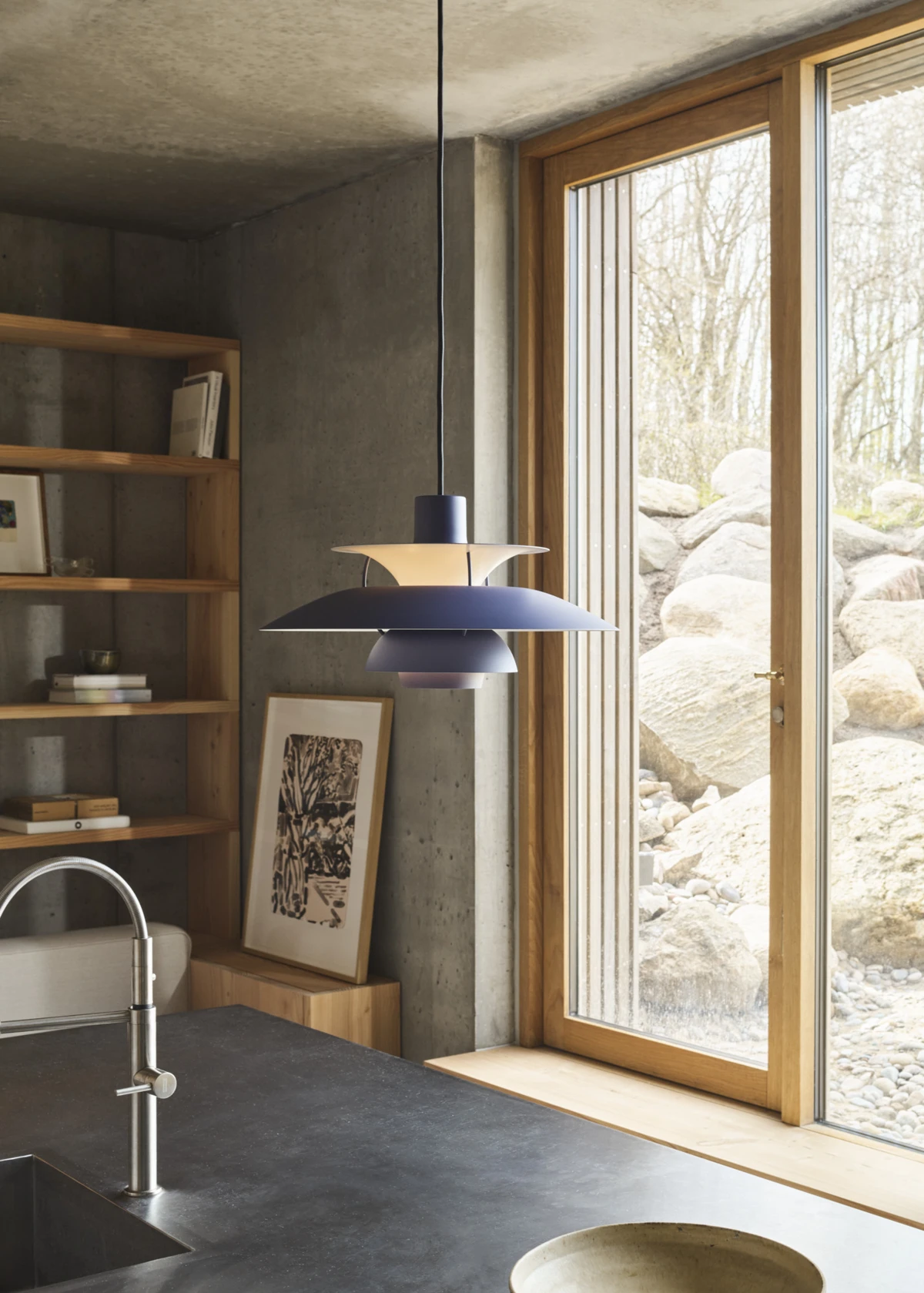 An indigo blue PH 5 pendant hangs above a kitchen island in a room with concrete walls and wood details. © 2026 Louis Poulsen.