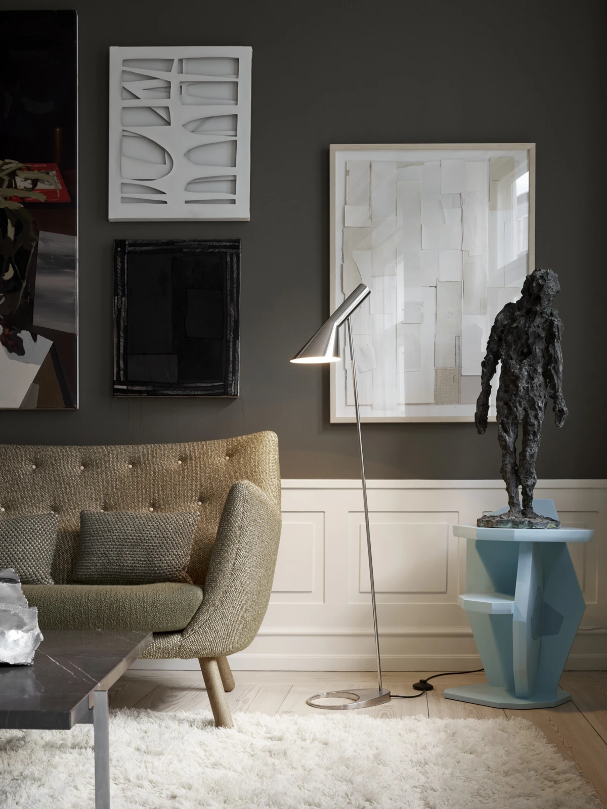 A metal AJ Floor Lamp casts light beside an upholstered sofa in an art-filled living room. © 2026 Louis Poulsen.