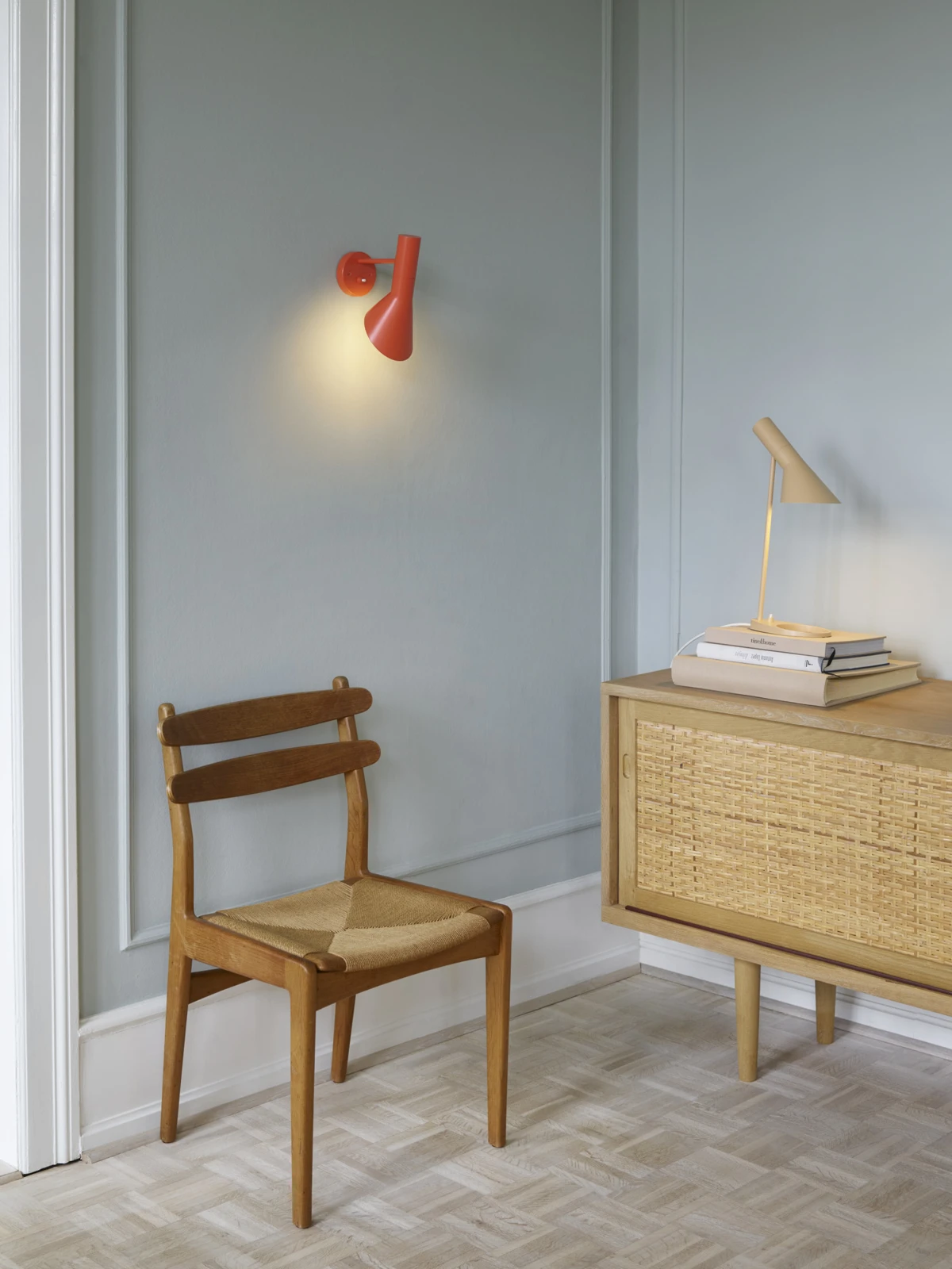 An orange AJ Wall Lamp lights the chair, while a Warm Sand AJ Mini Table Lamp sits on a rattan-fronted cabinet. © 2026 Louis Poulsen.