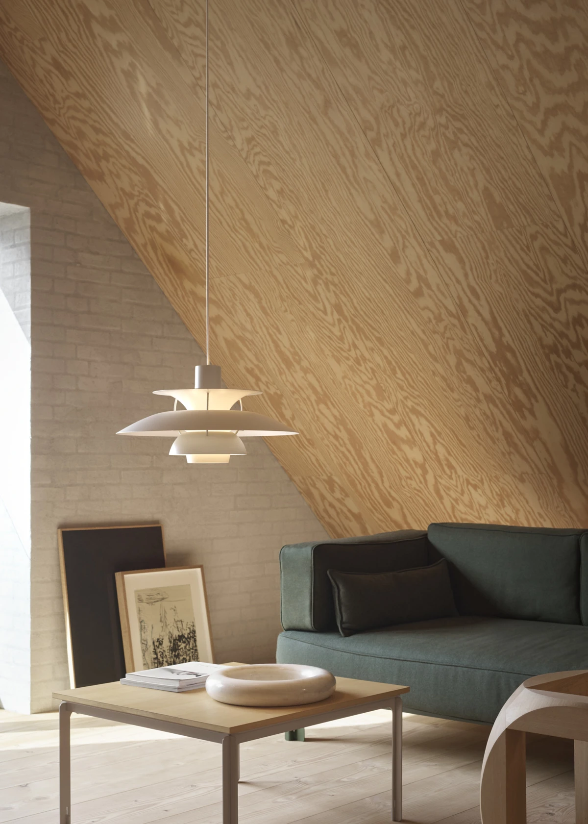 Een PH 5 hanglamp in licht pewter hangt boven een salontafel in een woonkamer met een houten schuin plafond en een bakstenen muur. © 2026 Louis Poulsen.