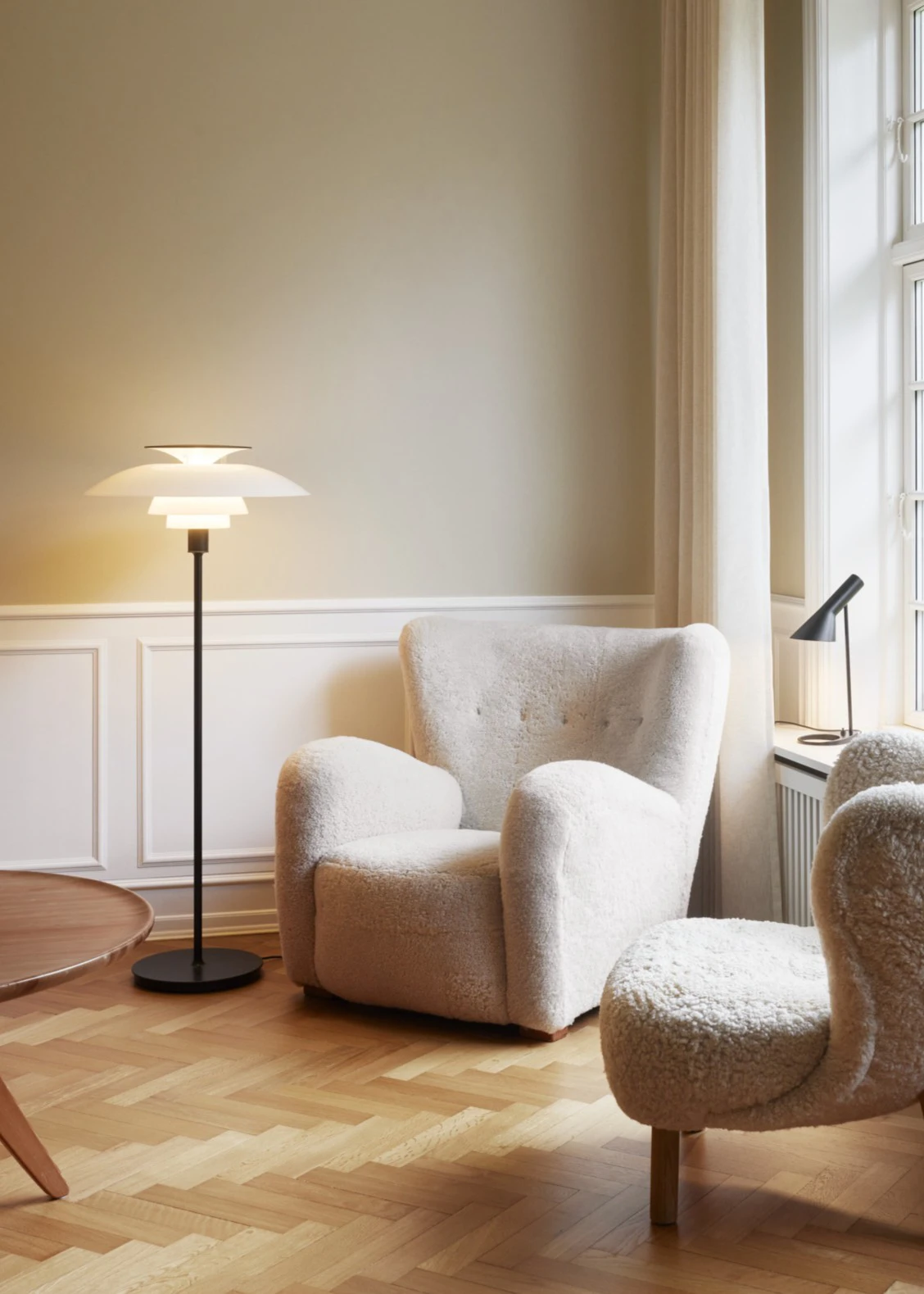 A white PH 80 Floor Lamp and a black AJ Mini Table Lamp in a bright living room with upholstered armchairs. © 2026 Louis Poulsen.