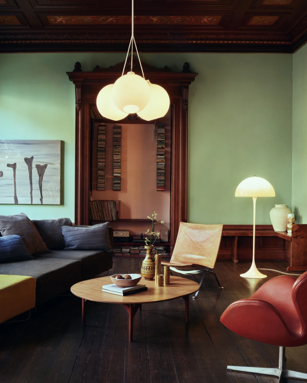 A Wohlert Pendant and Panthella Floor Lamp illuminate a living room with dark wood panelling and green walls. © 2026 Louis Poulsen.