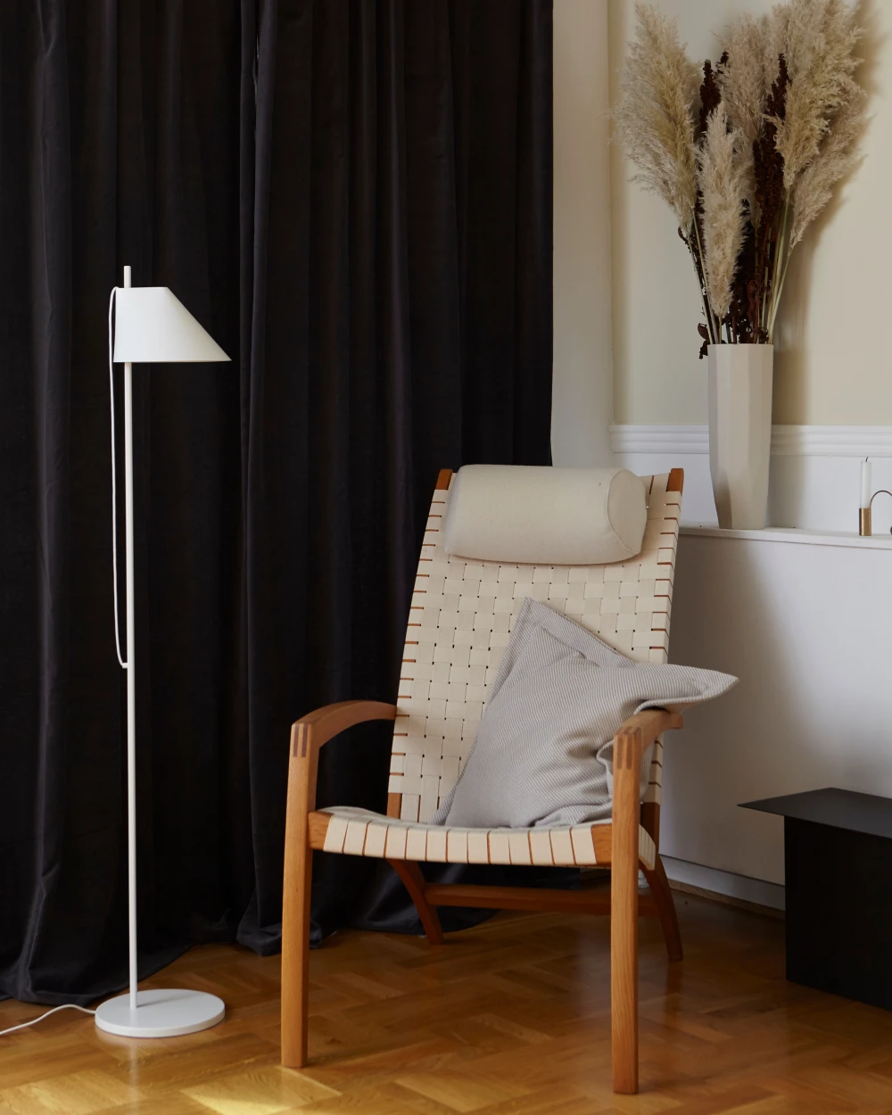 A white Yuh Floor Lamp stands beside a woven armchair in a living room with dark curtains and a wood floor. © 2026 Louis Poulsen.