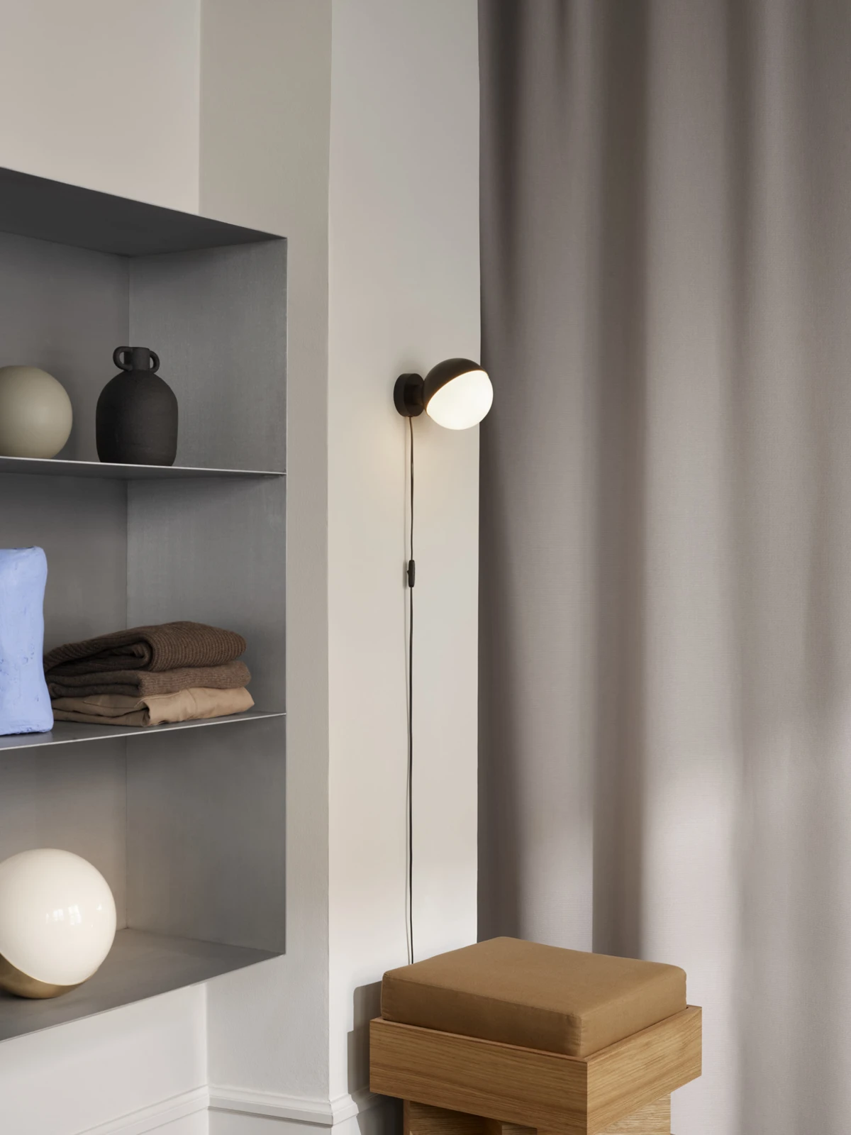 A black VL Studio Wall Lamp glows on a pale wall beside built-in shelves and a wooden bench. © 2026 Louis Poulsen.