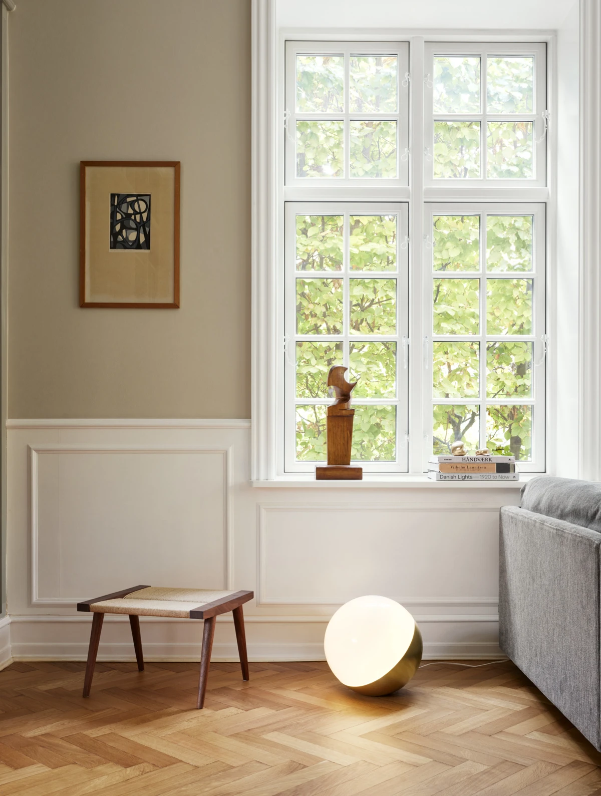 A brass VL Studio Table/Floor Lamp glows on the herringbone floor beside a grey sofa in a bright living room. © 2026 Louis Poulsen.
