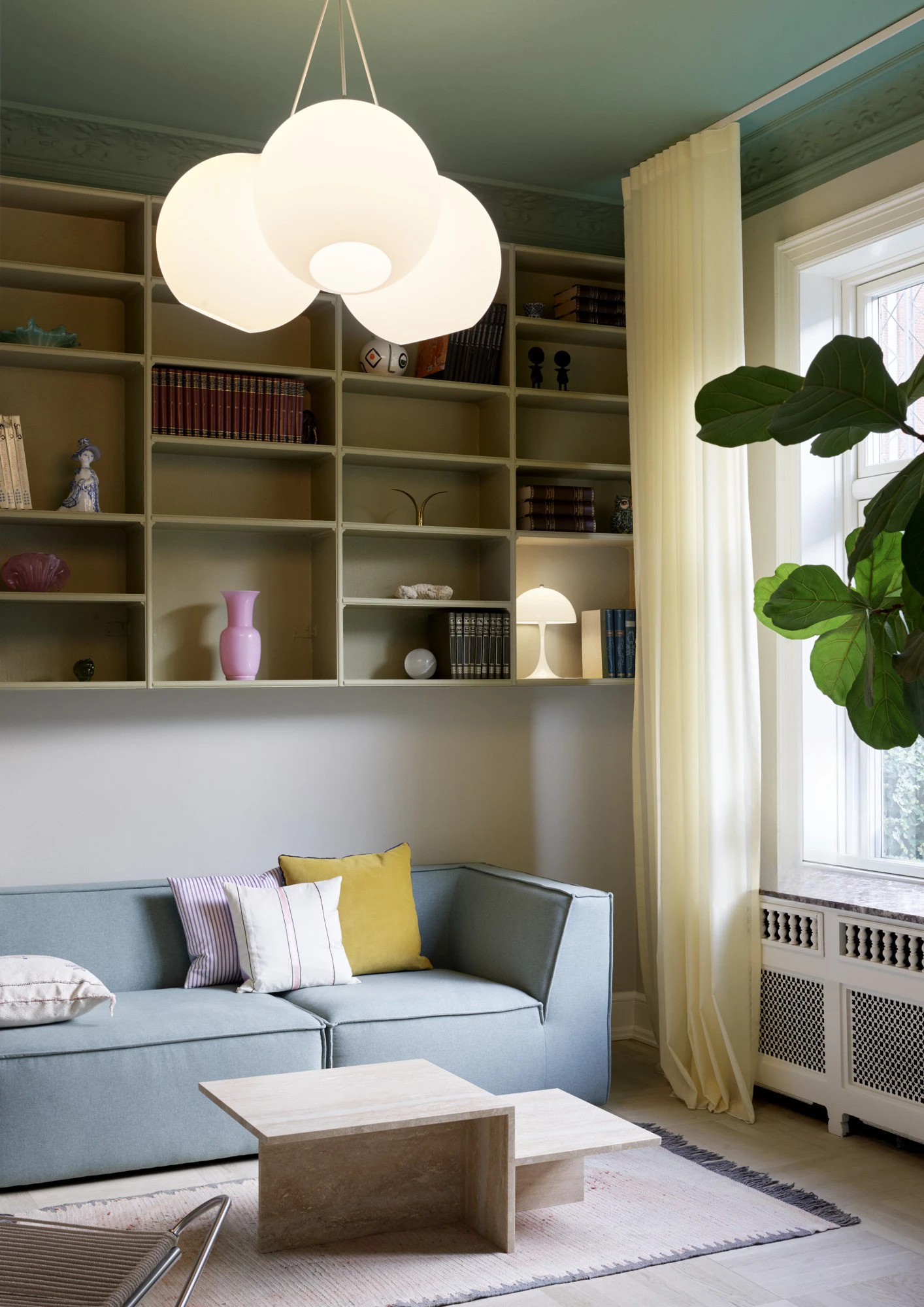 Een Wohlert hanglamp en Panthella 320 tafellamp verlichten een woonkamer met ingebouwde boekenkasten en een blauwe bank. © 2026 Louis Poulsen.