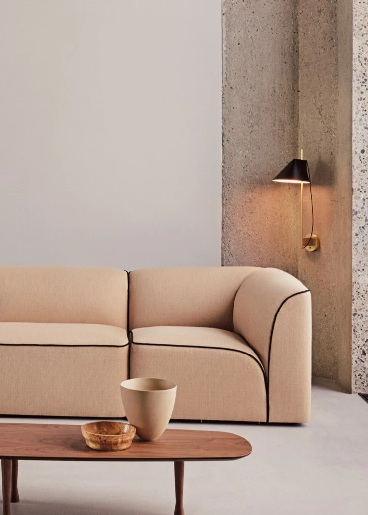 A brass Yuh Wall Lamp with a black shade casts warm light beside a sofa against a concrete wall. © 2026 Louis Poulsen.
