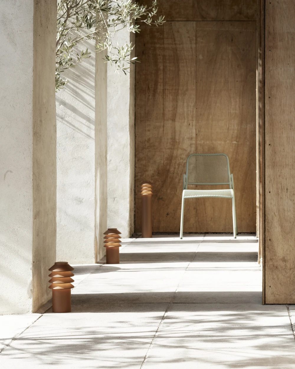 Copper-finish Bysted Garden Bollards line a sunlit patio walkway with stone paving and a wooden door. © 2026 Louis Poulsen.