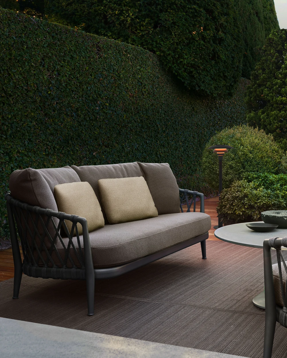 A PH 3-2½ Bollard glows beside a sofa on a patio surrounded by tall, leafy hedges. © 2026 Louis Poulsen.