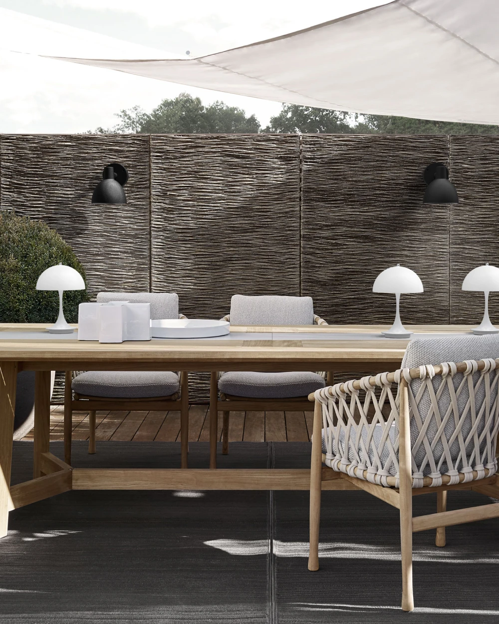Two black Toldbod 155 Outdoor Wall Lamps and white Panthella 160 Portable Lamps on a patio beside a wooden dining table. © 2026 Louis Poulsen.