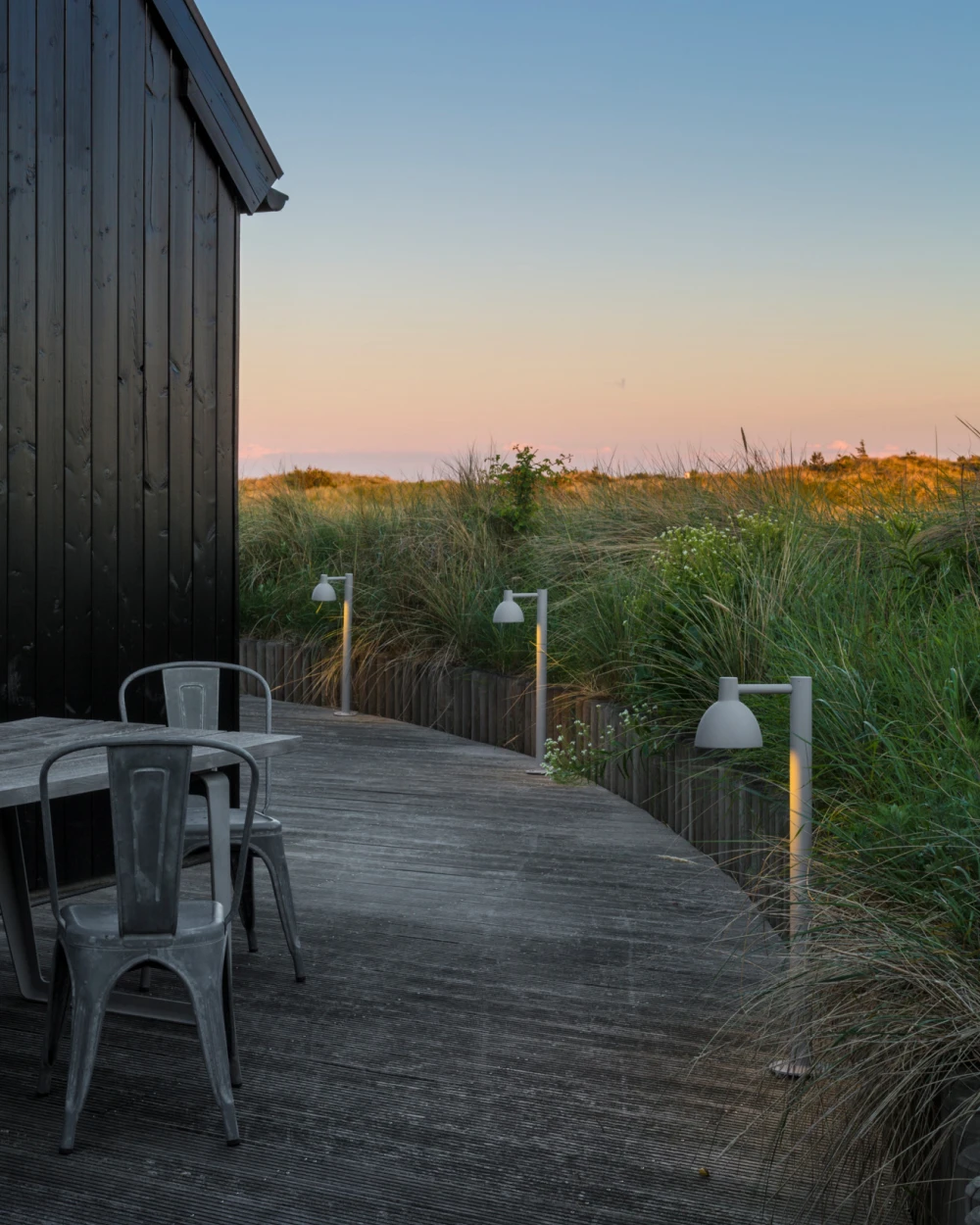 Grey Toldbod 155 Bollards light a timber deck beside dune grasses at dusk. © 2026 Louis Poulsen.