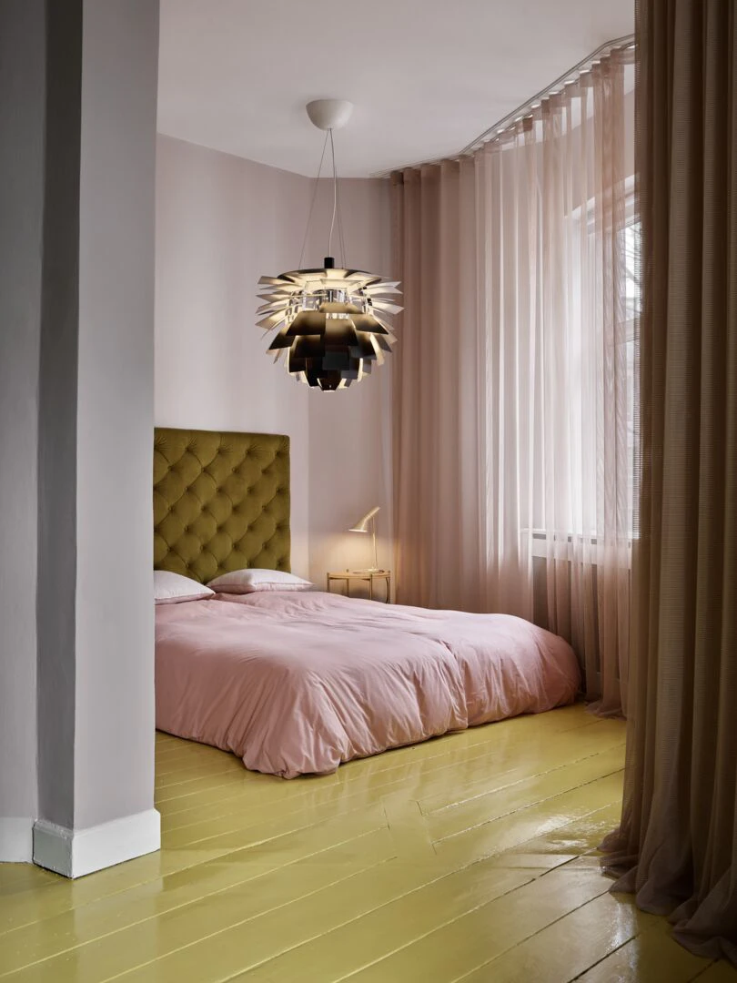 En PH Artichoke-pendel hänger över en säng med rosa sängkläder, olivgrön sänggavel och gulaktigt trägolv. © 2026 Louis Poulsen.
