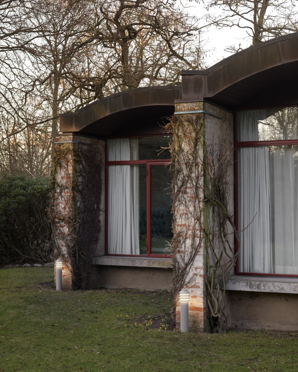 Two Bysted Garden Bollards stand beside a modern brick house in a garden with bare trees. © 2026 Louis Poulsen.