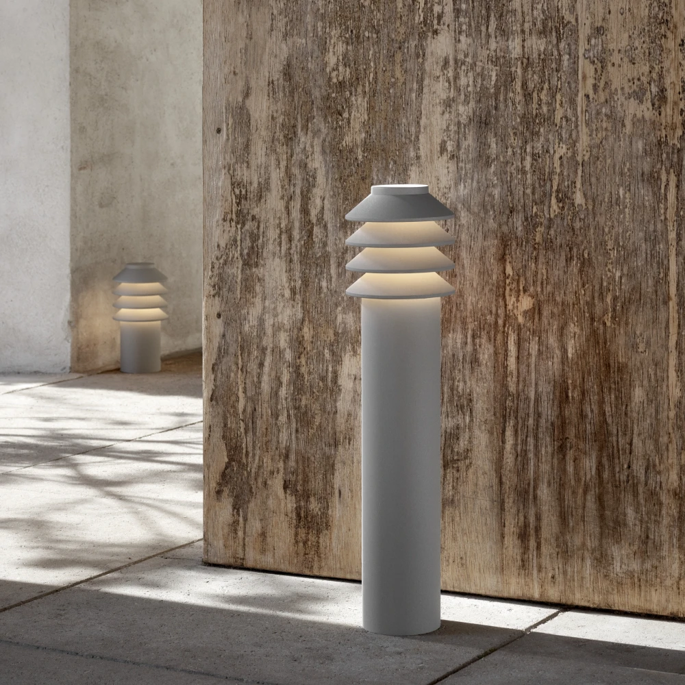 A grey Bysted Garden Bollard with glowing layered shades beside a weathered wooden wall on a paved terrace. © 2026 Louis Poulsen.