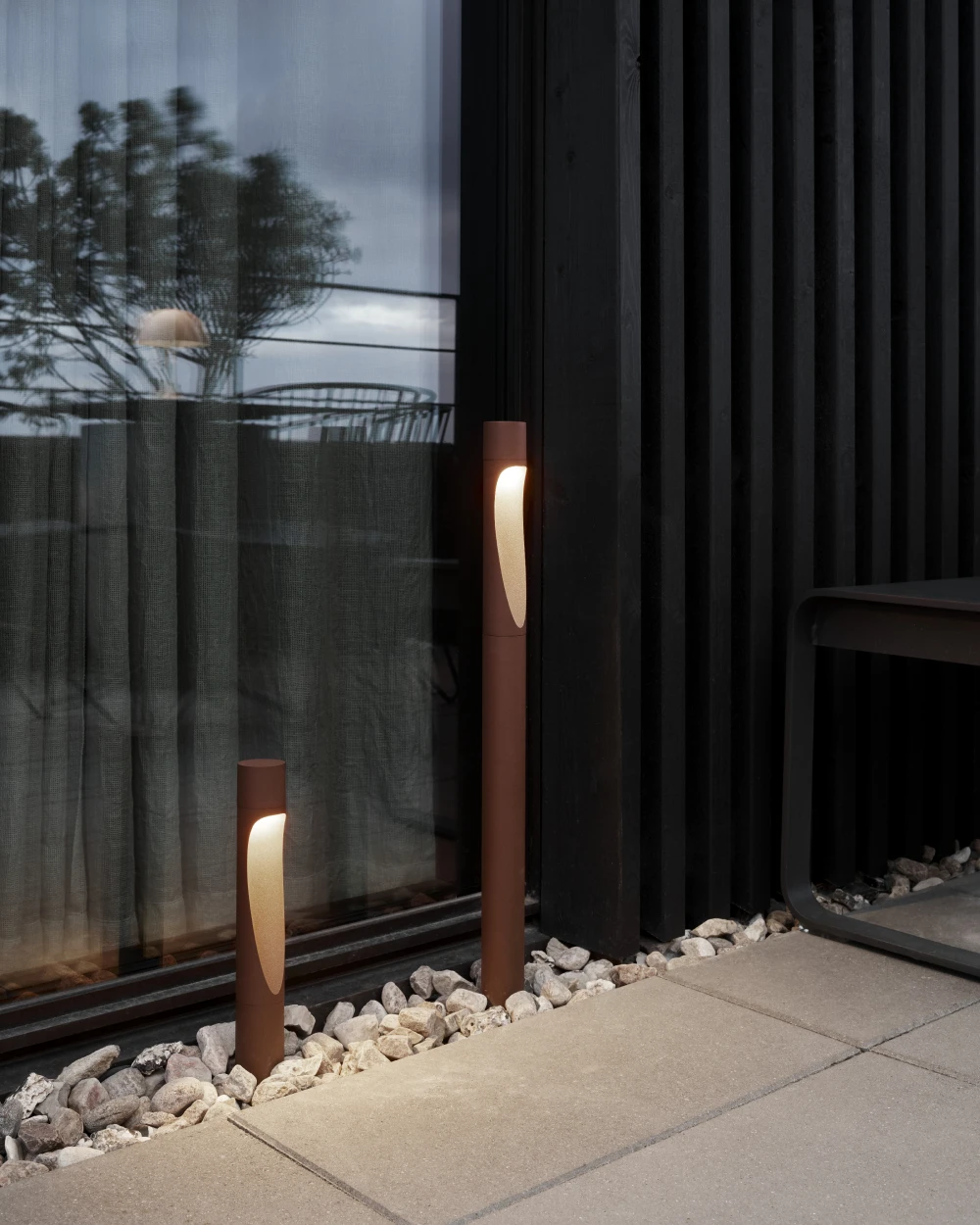 Two brown Flindt Garden Bollards illuminate a strip of stones beside a modern home. © 2026 Louis Poulsen.