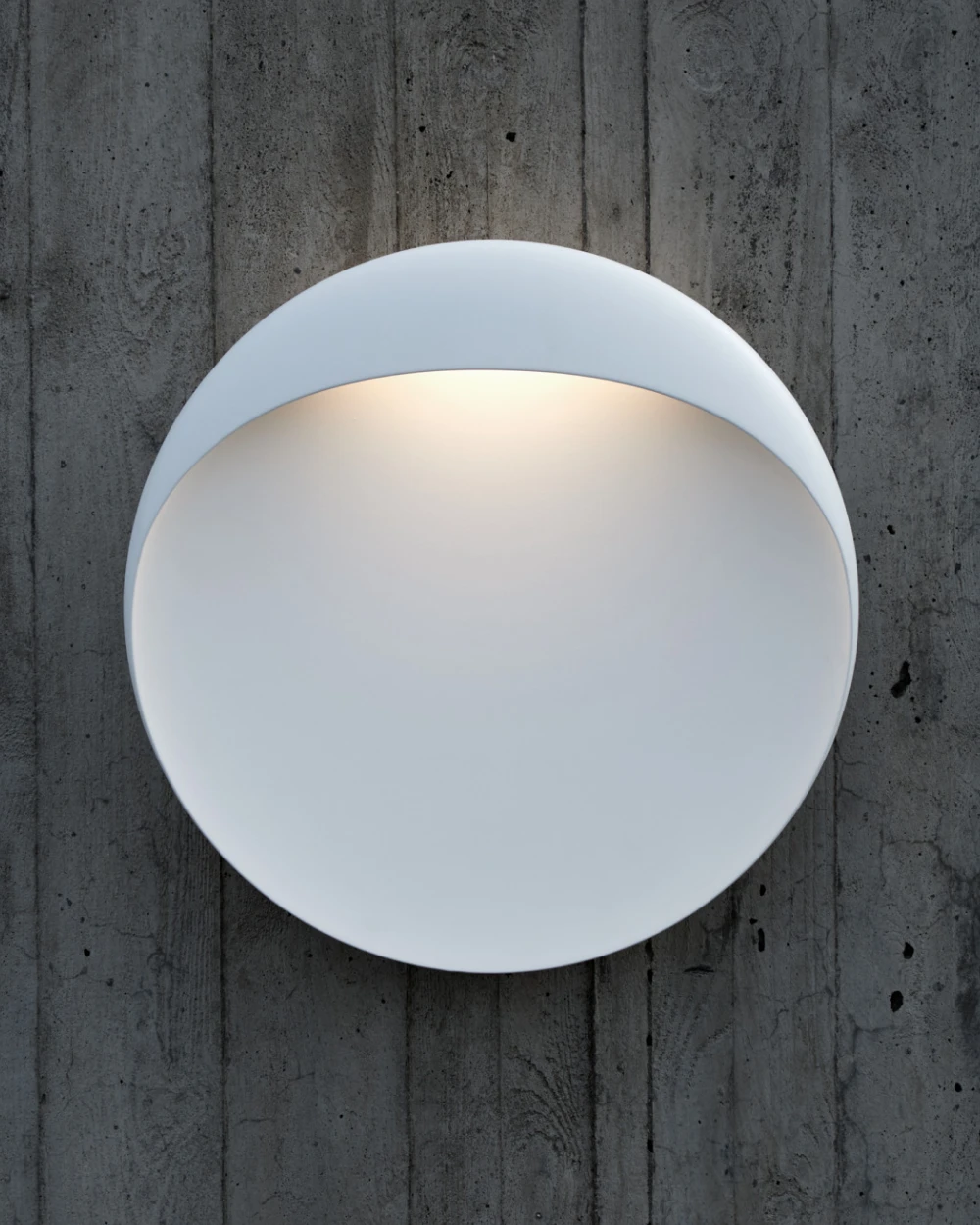 A white Flindt Wall Lamp casts a warm glow on a dark concrete wall outdoors. © 2026 Louis Poulsen.