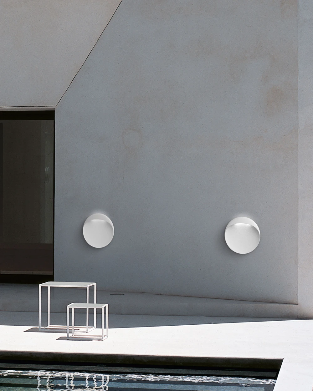 Two white Flindt Wall Lamp wall lights illuminate a concrete wall beside an outdoor pool. © 2026 Louis Poulsen.