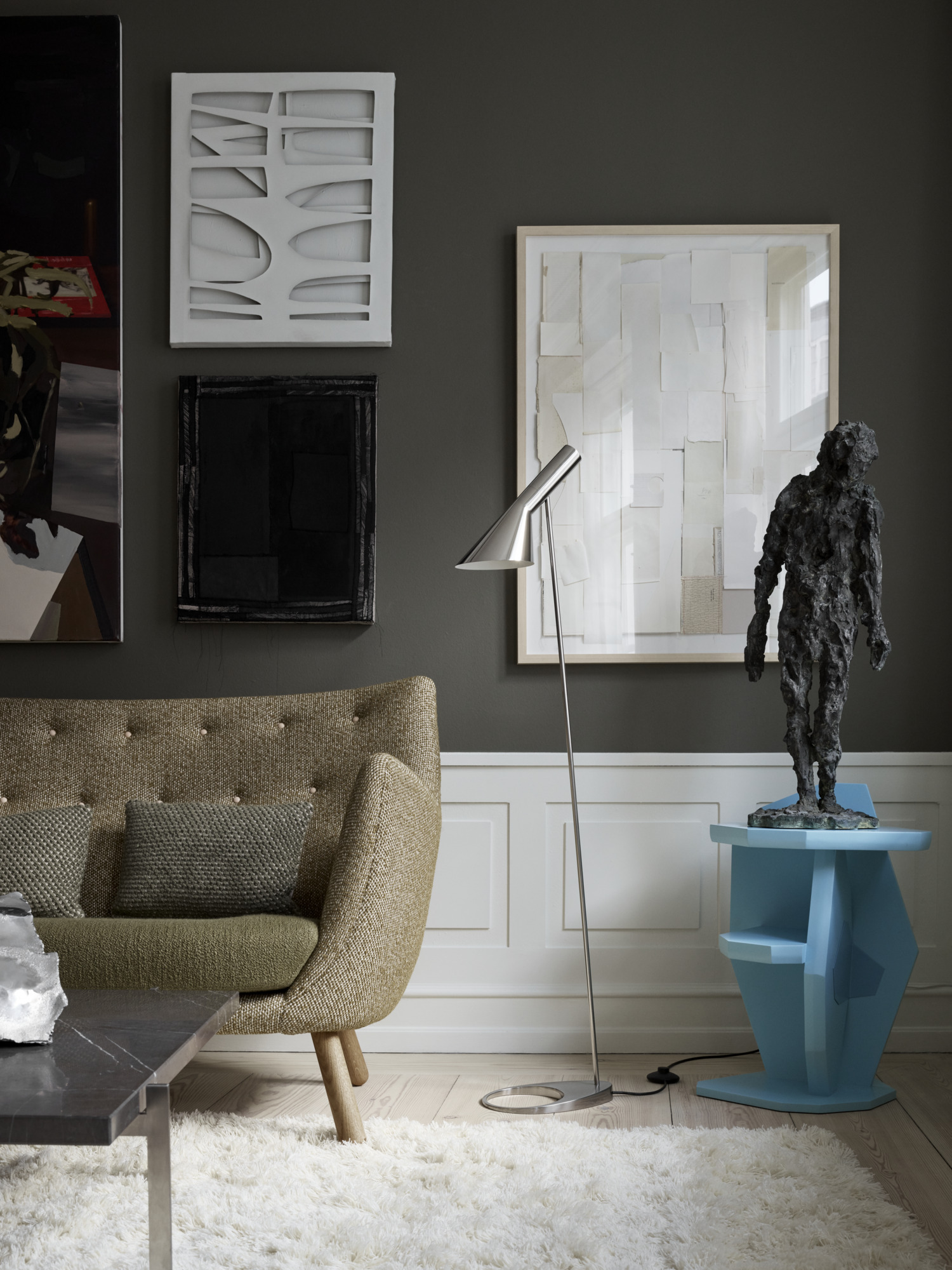 A silver-toned AJ Floor Lamp stands beside a textured sofa in a dark living room filled with artwork. © 2026 Louis Poulsen.