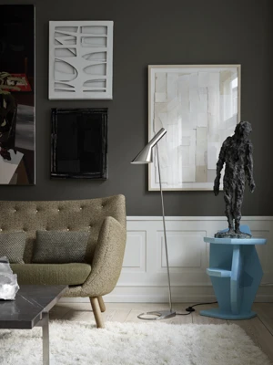 A silver-toned AJ Floor Lamp stands beside a textured sofa in a dark living room filled with artwork. © 2026 Louis Poulsen.