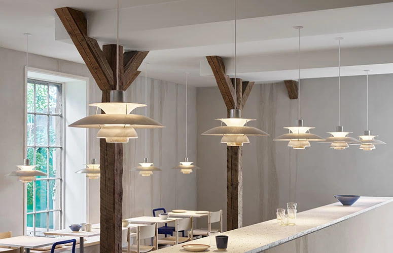 Light-coloured PH 5 Retake pendants hang above a long kitchen island and dining tables beneath exposed wooden beams. © 2026 Louis Poulsen.