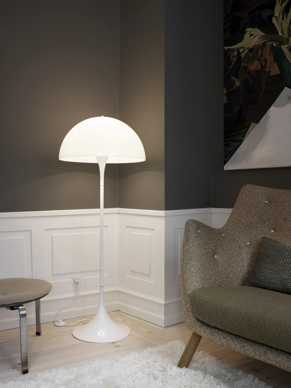 Een witte Panthella vloerlamp verlicht een hoek naast een fauteuil in een kamer met donkere muren en lichte lambrisering. © 2026 Louis Poulsen.