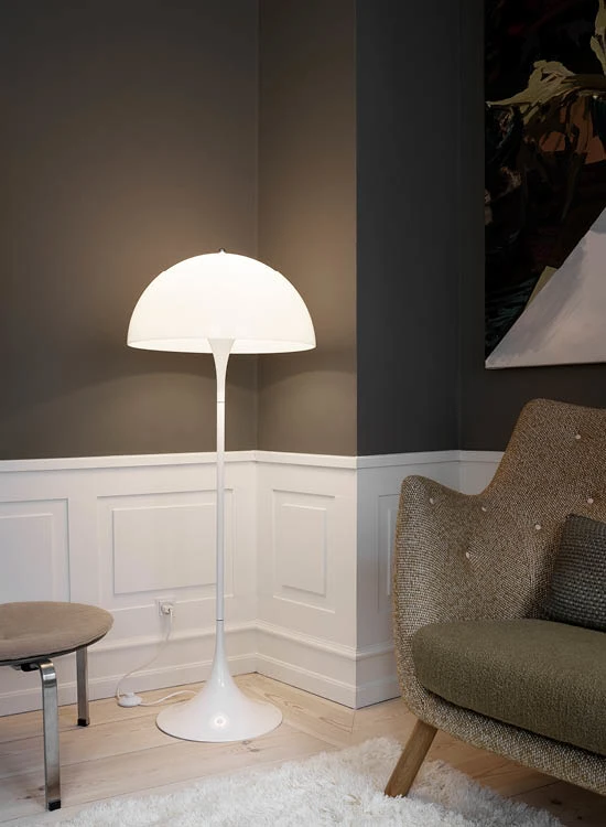 Hvid Panthella gulvlampe lyser varmt i et hjørne med panelvægge, trægulv og en polstret lænestol. © 2026 Louis Poulsen.