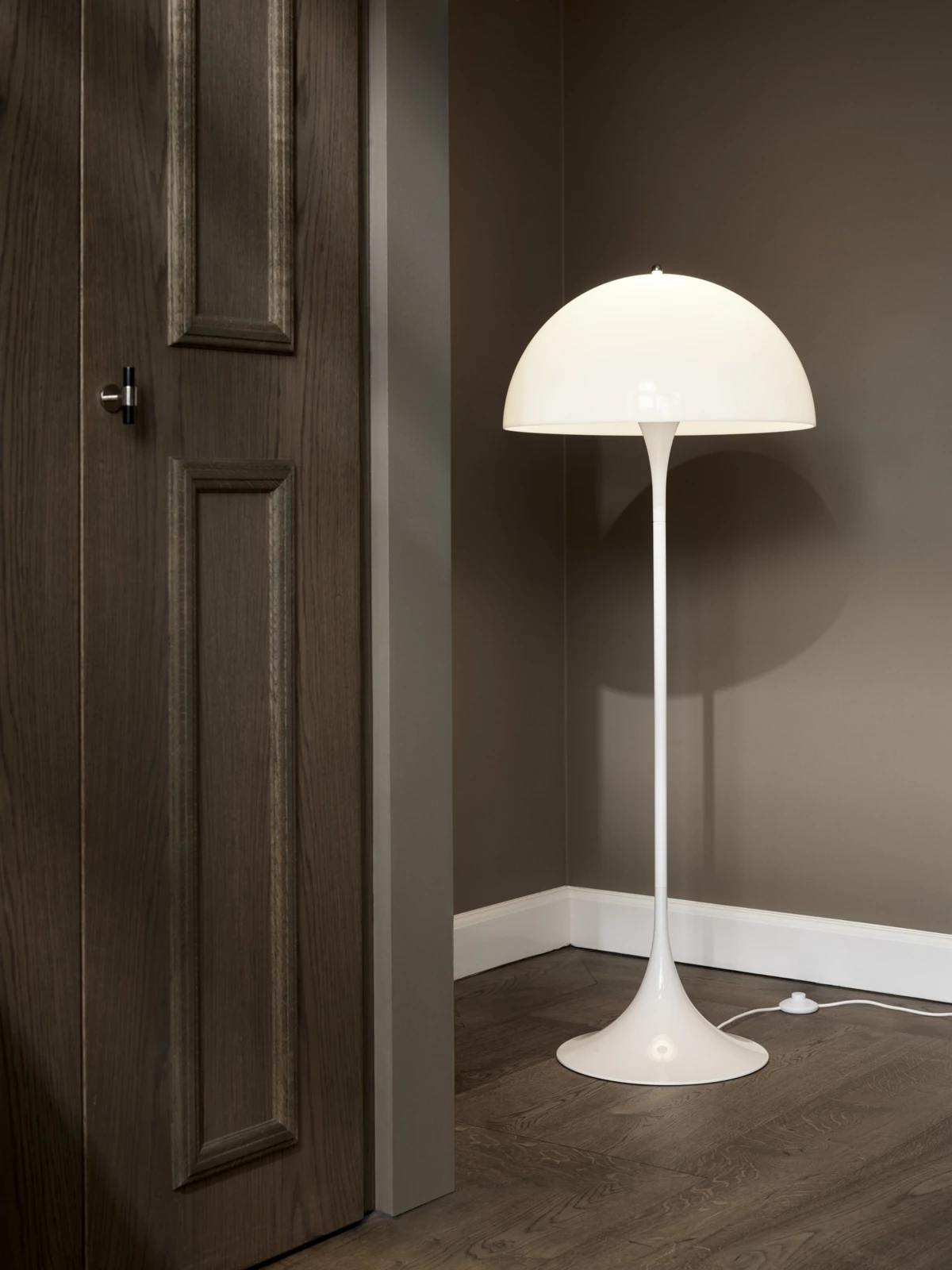 Een witte Panthella vloerlamp staat naast een donkere houten deur op de houten vloer van een ingetogen kamer. © 2026 Louis Poulsen.