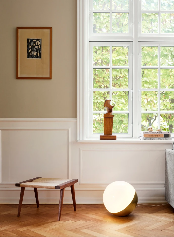 A VL Studio Table/Floor Lamp with a glowing white diffuser sits on herringbone flooring beside a large window. © 2026 Louis Poulsen.