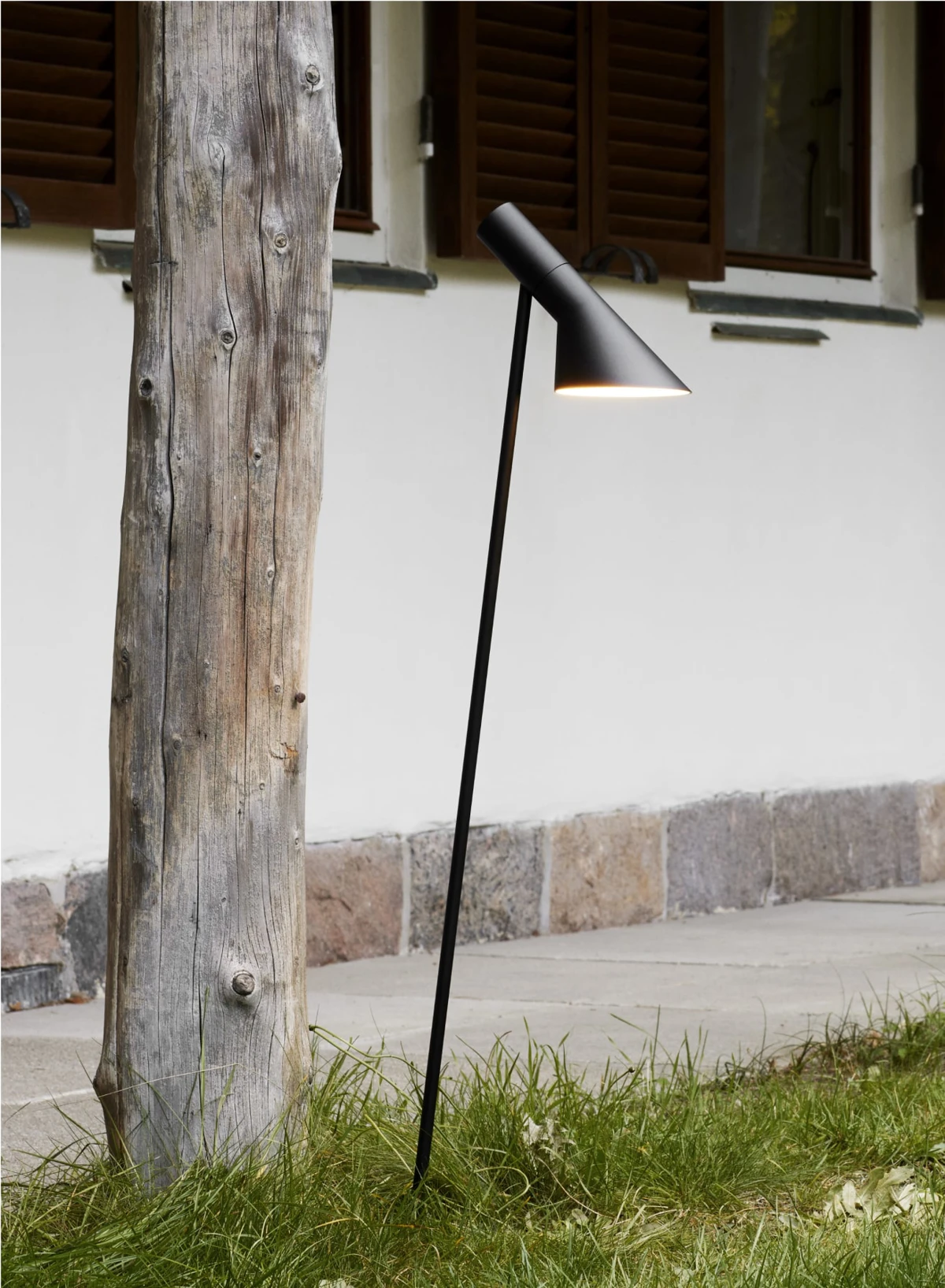 Een zwarte AJ Garden Bollard staat in het gras bij een wit huis met houten luiken. © 2026 Louis Poulsen.