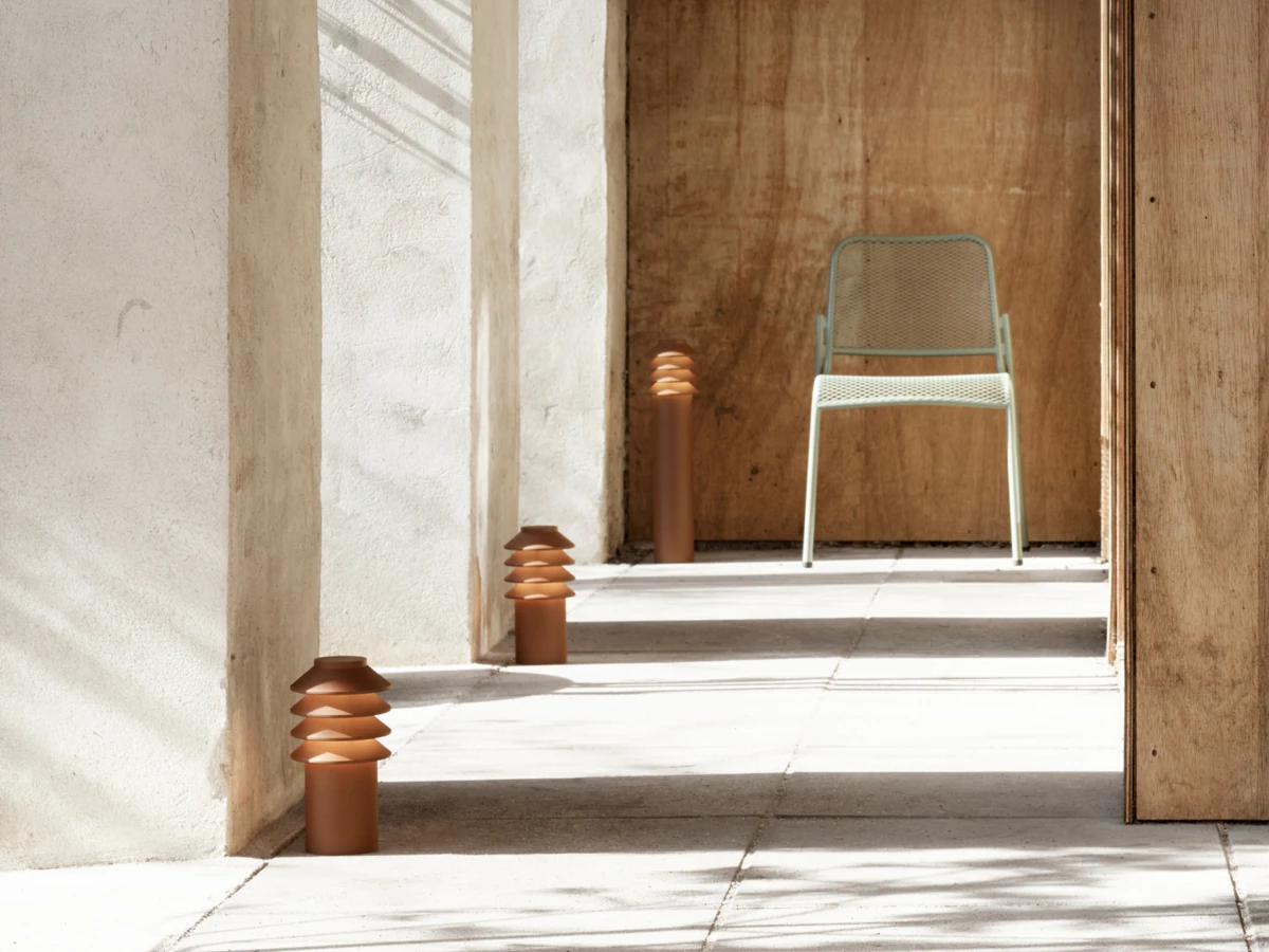 Rust-brown Bysted Garden Bollards line a sunlit walkway with pale stone walls and a wooden doorway. © 2026 Louis Poulsen.